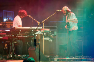 Two musicians perform on a dimly lit stage: one at a Roland keyboard with a mic over the stand, the other playing an electric guitar while smiling at the audience; colorful stage fog swirls around them.
