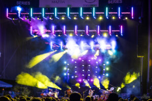 Outdoor concert stage bathed in multicolored lights and smoke as a band performs, with an audience in the foreground and Mazda Series banners overhead.