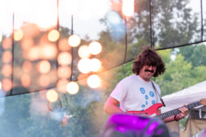 Male guitarist with shaggy brown hair and mustache, wearing a white T-shirt, playing a red electric guitar on an outdoor stage during a festival.