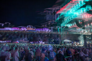 Crowd at a night concert with a neon-lit stage and colorful light trails above the audience
