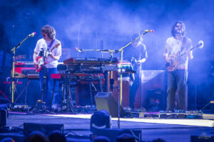 Three musicians performing on a foggy, blue-lit stage with keyboards and guitars and pedalboards at their feet.
