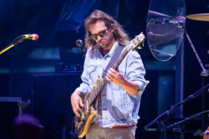 Musician with long hair and sunglasses playing a bass guitar on a dimly lit stage in a denim jacket.