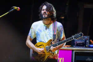 A bearded guitarist with a tie-dye shirt playing a sunburst semi-hollow electric guitar on stage, microphone to his left and laptop rig in the background.