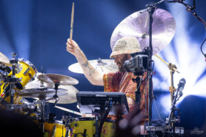 Drummer wearing a woven hat and patterned shirt raises a drumstick while playing a bright yellow drum set on stage.