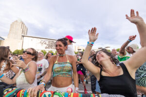 Crowd of people dancing and raising hands at an outdoor festival with a city skyline in the background. (informative)