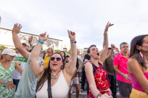 Crowd of diverse festival-goers cheering with arms raised at an outdoor event in daylight.