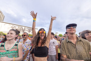 Woman in a black crop top and mirrored sunglasses raises her arms in excitement at a crowded outdoor event.",