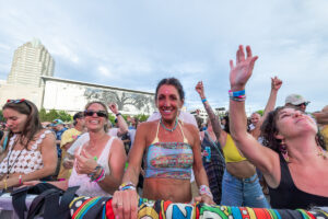 Festival crowd enjoying a concert; woman in a colorful halter top center smiles as she raises her hands.