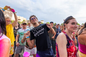 Crowd of people dancing at an outdoor concert, a man in sunglasses center-stage posing with friends, wearing a black T-shirt and colorful bracelets while others wear bright, tie-dye clothing.