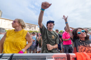 Crowd at an outdoor concert dancing with hands in the air; central man in a green shirt and blue cap raises his fist.