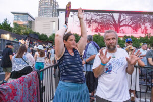 Woman in striped tank top raises a colorful item overhead at a crowded outdoor event; man nearby looks surprised with hands up.