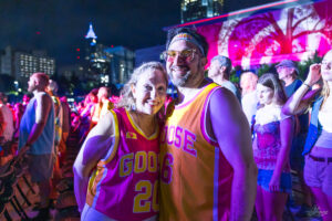 Smiling couple in bright pink and orange basketball jerseys posing together at a nighttime outdoor event, with a cheering crowd and city lights in the background.