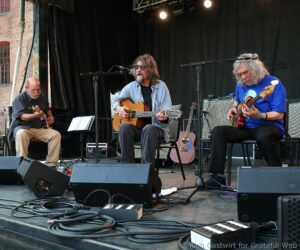 Three musicians on a stage playing guitars, with microphones, amps, and tangled cables surrounding them.