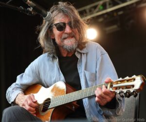 Older man with gray hair and beard wearing sunglasses, playing an acoustic guitar on stage with a microphone overhead.