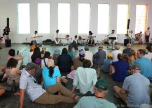 Wide view of a live band performing in a bright, minimalist hall while a large crowd sits on the floor watching the show.