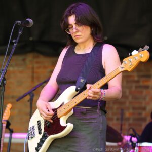 Female bassist playing a white Fender bass on stage, focused, wearing glasses and a dark sleeveless top.