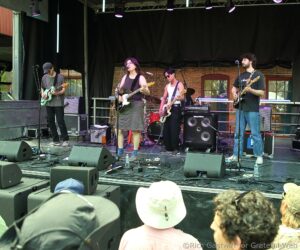 A four‑member band performs on an outdoor stage with guitars and microphones, brick backdrop and audience in the foreground coupled with stage equipment.