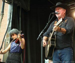Guitarist wearing a black hat sings into a microphone during a live stage performance, with a violinist visible beside him.