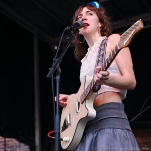 Female guitarist with red hair singing into a microphone on stage while playing an electric guitar on a dark background.