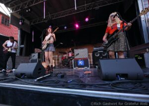 Band performing on an outdoor stage: guitarist left, singer center with guitar, bassist right in red shirt, drummer in back