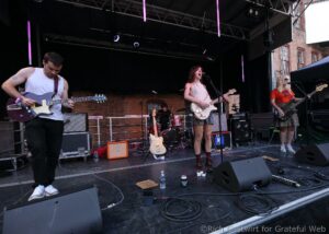 A rock band performs on an outdoor stage: guitarist in white tank top left, female vocalist with cream guitar center, drummer behind them, bassist on the right, brick building backdrop and stage gear around.