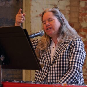 Woman with long gray hair wearing a black-and-white plaid shirt sings into a microphone at a keyboard in a brick-walled venue.