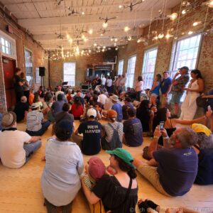 Audience seated on the wooden floor of a brick-walled venue, watching a live performance under hanging light bulbs. (Informative: interior concert/hall ambience)