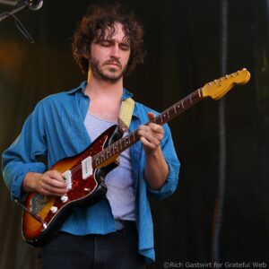 Male guitarist performing on stage with a sunburst electric guitar and blue striped shirt, close-up of his torso and instrument neck against a dark backdrop.