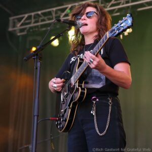 Female guitarist singing into a microphone on stage, wearing sunglasses and a black t-shirt, with an electric guitar and accessories visible in a close-up shot.