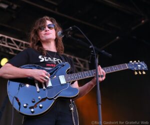 Female musician wearing sunglasses, playing a blue electric guitar on stage with a nearby microphone.",