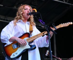 Female guitarist singing into a microphone on stage while playing a sunburst Fender Telecaster guitar with white shirt and curly blonde hair.