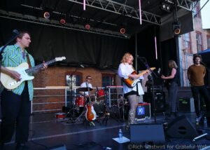 A rock band performing on an outdoor stage: guitarist in a striped shirt left, female guitarist sings center, drummer in back, brick building backdrop and stage lights above.