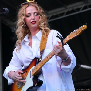 Female musician playing an orange-electric guitar on stage, looking down at the fretboard in a white button-down shirt.