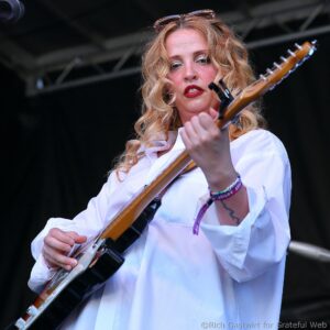 Female guitarist in a white shirt playing an electric guitar on stage, looking toward the audience/camera with red lipstick.