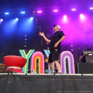 Male performer on stage wearing a white cap and sunglasses, reaching toward a colorful rainbow backdrop under blue and purple lights.