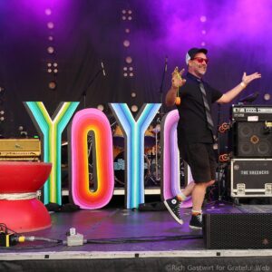 Male performer on a colorful stage, wearing a cap and red sunglasses, speaking or singing with arms outstretched; large rainbow letters spell a word in front.