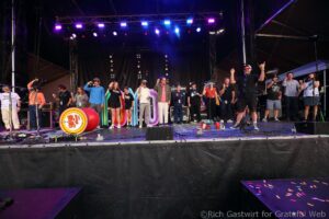 Group of people on a stage at a festival, posing for a photo with colorful rainbow décor and bright stage lights.