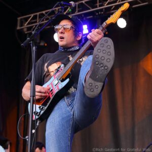Guitarist performing on stage, wearing sunglasses and a black cap, playing an electric guitar with a microphone nearby.