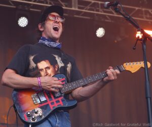 Guitarist singing into a microphone on stage, wearing a black cap, sunglasses, and bandana, playing a sunburst electric guitar.