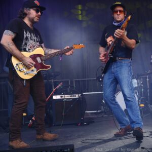 Two guitarists on a stage playing electric guitars; left musician with tan Tele-style guitar, black shirt, hat and tattoos; right musician with darker guitar, jeans, bandana, sunglasses.