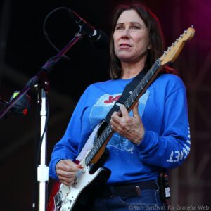 Woman guitarist on stage, singing into a microphone while playing a red-and-white electric guitar.