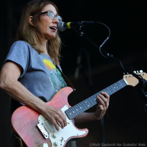 Woman singing into a microphone while playing a pink electric guitar on stage; wearing glasses and a gray T-shirt.