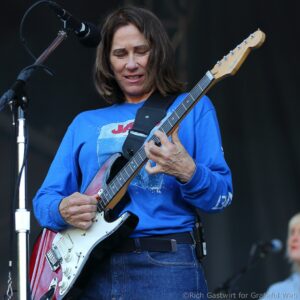 Guitarist in a blue shirt playing a red Fender Stratocaster on a live stage, microphone in front.
