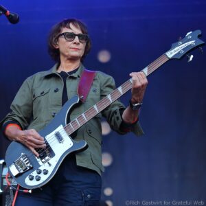 Musician wearing glasses and an olive-green jacket playing a blue bass guitar on stage at a concert.