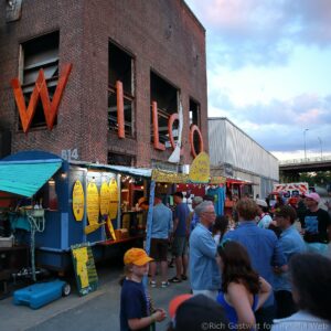 Crowd at an outdoor food market along a brick building with colorful signs and menu boards for dumplings and snacks.