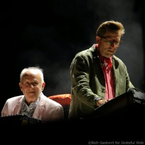 Two musicians on stage: an older man at a keyboard on the left and a younger man with glasses and a red scarf standing at a piano on the right, dark background