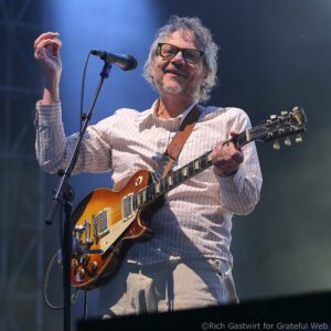 Male guitarist with gray hair and glasses playing a sunburst electric guitar on stage, singing into a microphone.