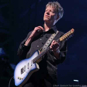 Male guitarist in a striped shirt playing a white/blue electric guitar on stage, close-up from the chest up at a live concert.