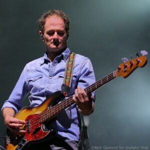 Male musician playing a sunburst bass guitar, wearing a blue denim shirt and patterned strap, eyes closed.