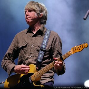 Guitarist wearing a brown shirt playing a yellow electric guitar on stage with a patterned blue strap visible at the shoulder.